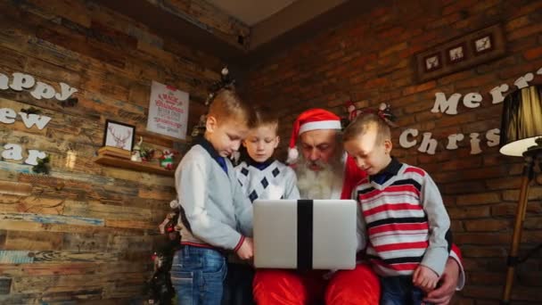 Добрые парни помогают Санта-Клаусу писать письма на ноутбуке в украшенной комнате — стоковое видео