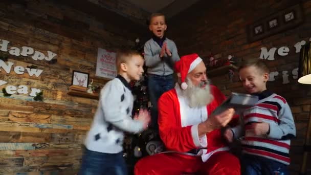 Санта-Клаус из трех мальчиков-помощников будет работать с гаджетом в украшенной к празднику комнате с красивой елкой — стоковое видео