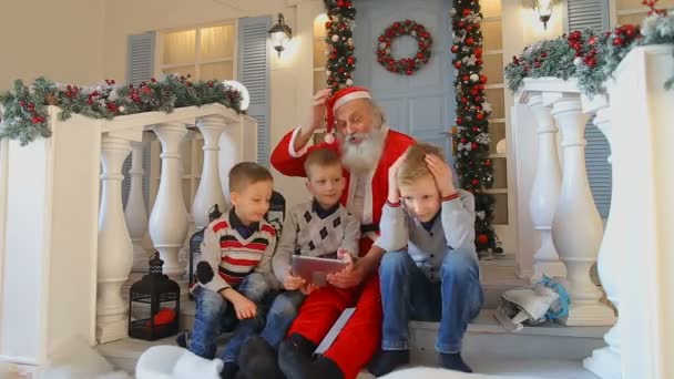 Санта Клаус і трьох братів проходять через втрати в грі планшетний ПК, сидячи на сходах на ганку будинку прикрашали свята — стокове відео