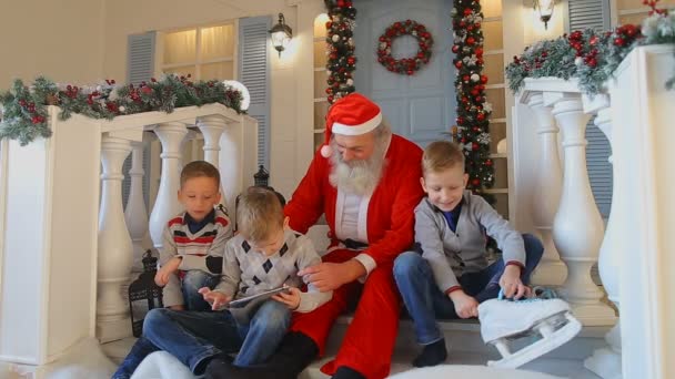 Przyjazny dla małych facetów aktywnie spędzają wolny czas z Santa siedzi na werandzie domu piękne ozdoby i bujny drzewo pokryte śniegiem na dziedzińcu — Wideo stockowe