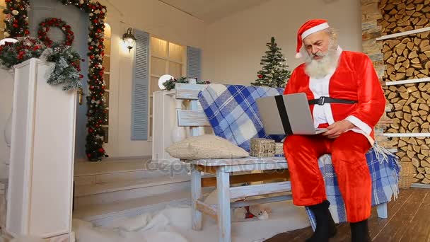 Santa aspirujących mówi na videocall z Boże Narodzenie elfy na laptopie siedząc na ławce w courtyard urządzone na wakacje — Wideo stockowe