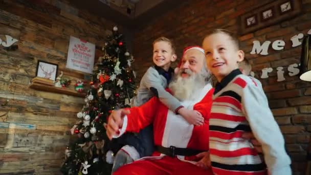 Drei glühende Jungen Brüder mit Spaß springen auf Weihnachtsmann claus im Zimmer geschmückten Weihnachtsbaum — Stockvideo