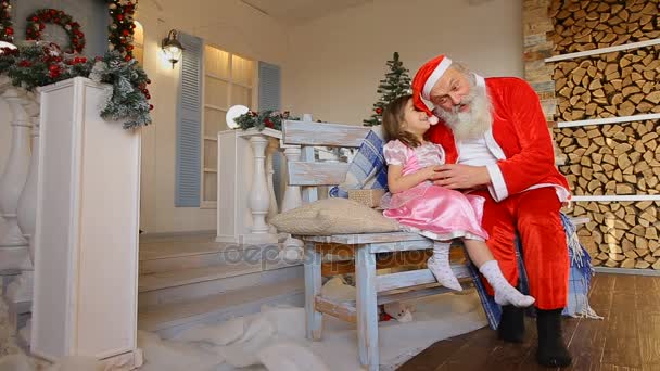 Mooi meisje fluistert Santa geheime wens, zittend naast op bankje in de binnenplaats die versierd met vakantie — Stockvideo