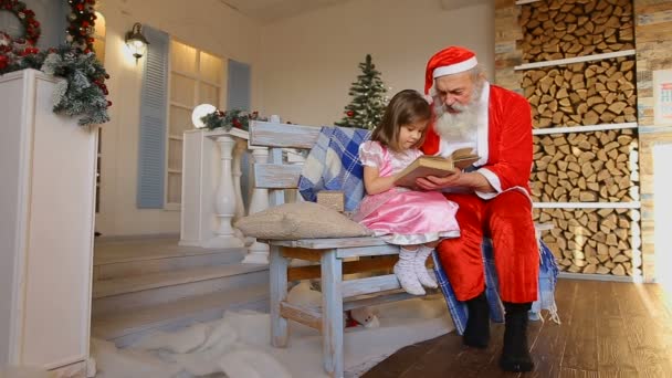 Nadšené dítě ženského pohlaví se dívá na jasné obrázky v knize spolu s Santa Claus na lavici poblíž krásně zdobený dům pro dovolenou — Stock video