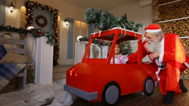 Дед Мороз подарил девочке большой игрушечный автомобиль на крыльце дома, украшенный к празднику — стоковое видео
