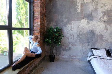 Çekici genç kadın gülümseyerek poz veriyor ve pencere pervazına üzerinde oturur