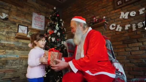 Маленькая девочка приходит к Санта-Клаусу, представляет новогодний подарок и обнимает дедушку Рождества — стоковое видео