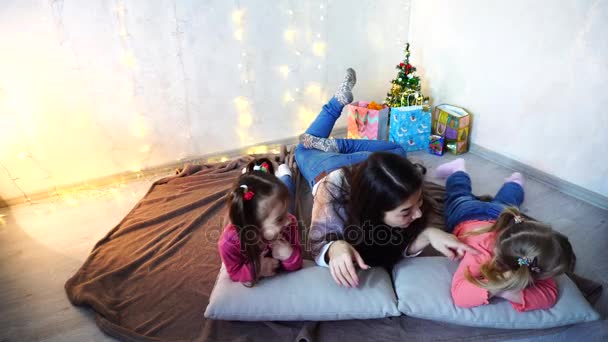 Schattig vrouwtje en twee kleine meisjes kinderen, poseren voor de camera en lacht, liggend op kussens, op de vloer in de kamer met garland en fir tree in avond. — Stockvideo