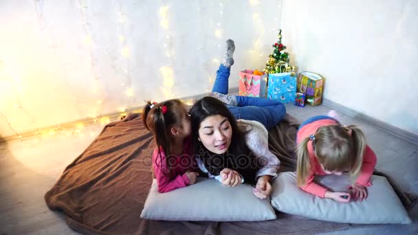 Linda hembra y dos niñas niños pequeños, posando para la cámara y sonriendo, acostado en cojines, en el suelo en la habitación con guirnalda y abeto en la noche . — Vídeo de stock