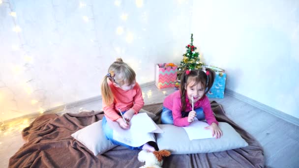 Lustige kleine Mädchen lachen und reden, posieren auf dem Boden liegend und auf Kissen an der Wand mit Girlanden und Christbaum am Tag. — Stockvideo