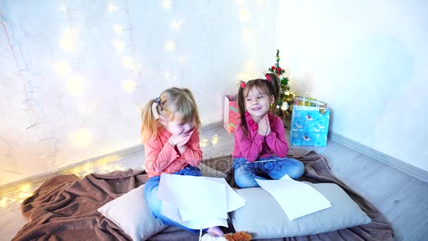 Små flickor skratta och prata, poserar liggande på golvet och på kuddar mot väggen med krans och julgran på dagtid. — Stockvideo