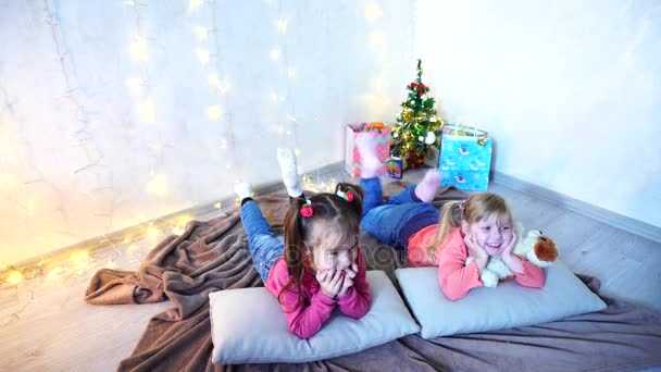 Маленькие девочки смеются и говорят, позируя лежа на полу и на подушках к стене с гирляндой и елкой в дневное время . — стоковое видео