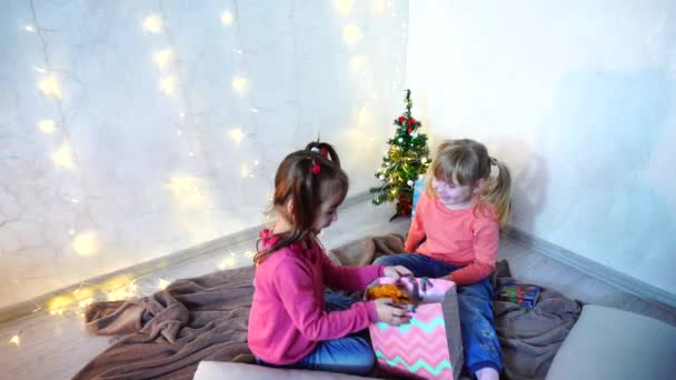 Actieve jonge kinderen van meisjes lachen en gek rond, zittend op de vloer en tapijt, tegen de achtergrond van de muur met garland en kerstboom in de middag. — Stockvideo