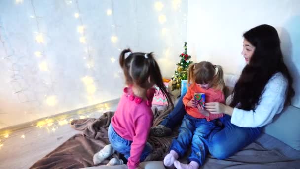 Vacker äldre syster att ha kul och leka med små flickor sitter på matta, mot bakgrund av julgran och en krans på väggen i sovrummet på tröskeln till jul. — Stockvideo