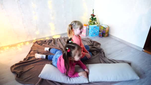 Lustige kleine Mädchen lachen und reden, posieren auf dem Boden liegend und auf Kissen an der Wand mit Girlanden und Christbaum am Tag. — Stockvideo