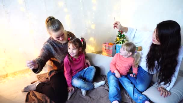 Weiblicher Schwestertanz mit weihnachtlichem Bengalfeuer am Boden sitzend im Raum auf dem Hintergrund der Wand mit Girlande und kleinem Baum. — Stockvideo