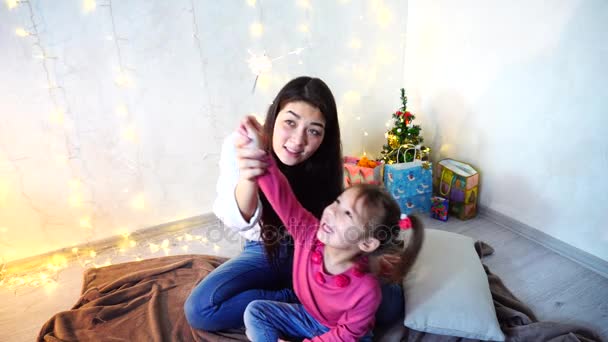 Communication de la sœur aînée avec la plus jeune fille qui passe du temps ensemble et assis sur le sol dans la chambre sur fond de mur avec guirlande et petit arbre . — Video