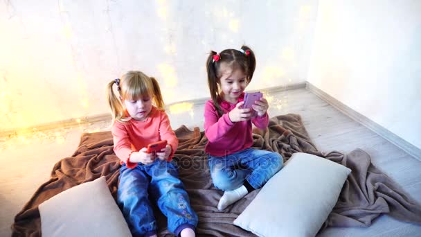 Happy μικρά κορίτσια χρησιμοποιούν smartphones για ψυχαγωγία και καθίστε στο πάτωμα σε φωτεινή αίθουσα με γιρλάντα στον τοίχο. — Αρχείο Βίντεο