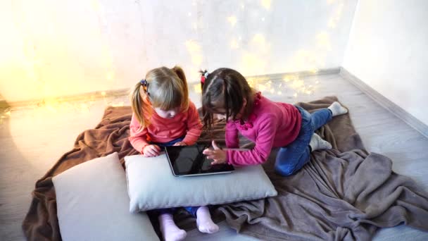 Meninas envolvidas no uso de tablet e sentar no chão em quarto brilhante com guirlanda na parede . — Vídeo de Stock