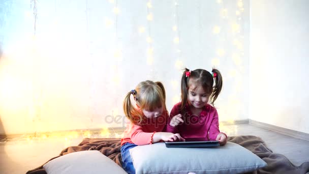 Pequenas meninas envolvidas no uso de tablet e sentar no chão em quarto brilhante com guirlanda na parede . — Vídeo de Stock