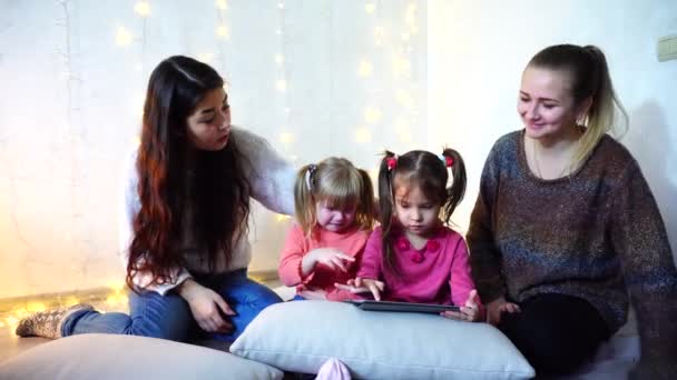 Oudere vrouwen en baby meisjes vier communiceren en bezig met eigen zaken, zittend op de vloer op de achtergrond van de muur met garland op kamer. — Stockvideo