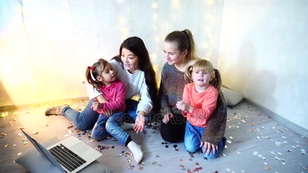 Ältere Frauen verbringen Zeit mit jüngeren Mädchen und Schwestern mit Laptop und sitzen auf dem Fußboden an der Wand mit Girlanden im Zimmer. — Stockvideo