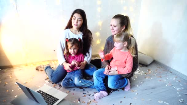 Zwei ältere Frauen verbringen Zeit mit jüngeren Mädchen und Schwestern mit Laptop und sitzen auf dem Boden vor der Wand mit Girlanden im Zimmer. — Stockvideo