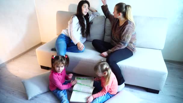 Duas jovens mães sentadas no sofá na sala de estar e crianças brincam no chão . — Vídeo de Stock