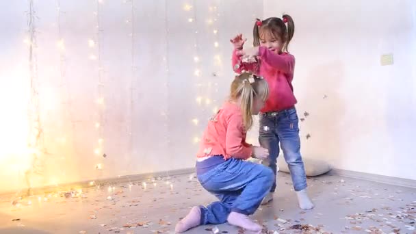 Twee meisjes spelen in woonkamer en pick-up kerst confetti van vloer. — Stockvideo