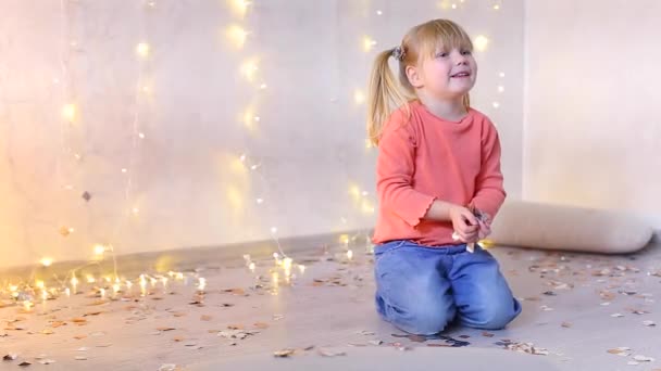 Niña rubia se sienta en el suelo en la sala de estar y vomita confeti brillante . — Vídeos de Stock