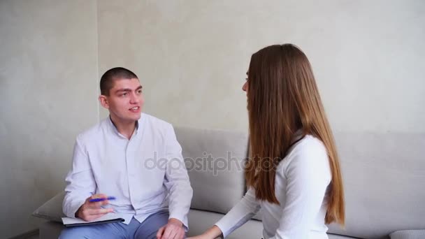 Mladá žena přišla k doktorovi, který naslouchá a píše. Guy a žena sedí v ordinaci na gauči na pozadí šedé stěny. — Stock video