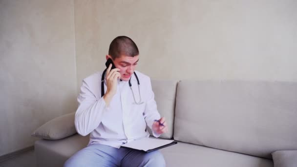 Doctor communicates on cell and negotiates with patient, sitting on couch in office of hospital. — Stock Video