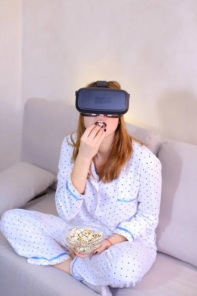 Cool girl watches video in VR glasses, sitting on couch in brigh