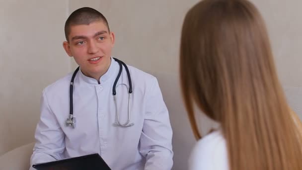 Homme médecin donne des instructions et des conseils à la fille à la réception dans le bureau médical, assis sur le canapé fond de mur gris . — Video