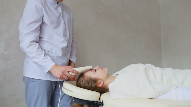 Мужчина-косметолог демонстрирует работу массажного аппарата для лица на фоне пациентки девушки, лежащей на диване в салоне . — стоковое видео