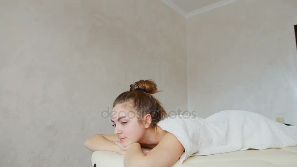 Όμορφη γυναίκα χαλαρώνει μετά το μασάζ και κοιτάζει προς τα κάτω, ξαπλωμένη στον καναπέ κάτω από την πετσέτα στο φωτεινό ιατρείο. — Αρχείο Βίντεο
