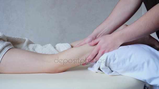 Профессиональный массажист разминает ноги пациентке, которая лежит на диване в светлом кабинете . — стоковое видео