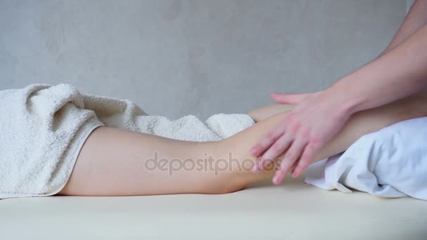 Профессиональный массажист разминает ноги пациентке, которая лежит на диване в светлом кабинете . — стоковое видео