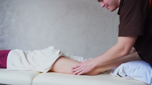 Professionelle Kerl Massagetherapeut von Hand macht Anti-Cellulite-Fußmassage weibliche Patientin, die auf der Couch im hellen Büro liegt. — Stockvideo