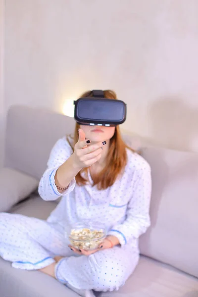 Cool girl watches video in VR glasses, sitting on couch in brigh