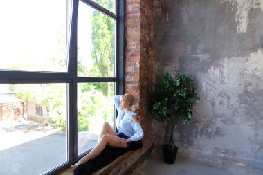 Çekici genç kadın gülümseyerek poz veriyor ve pencere pervazına üzerinde oturur