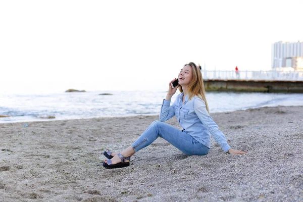 Beautiful female speaks on mobile with smile and sits on beach n — Stock Photo, Image