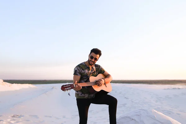 Handsome Arabian guy playing guitar, standing on hill among sand