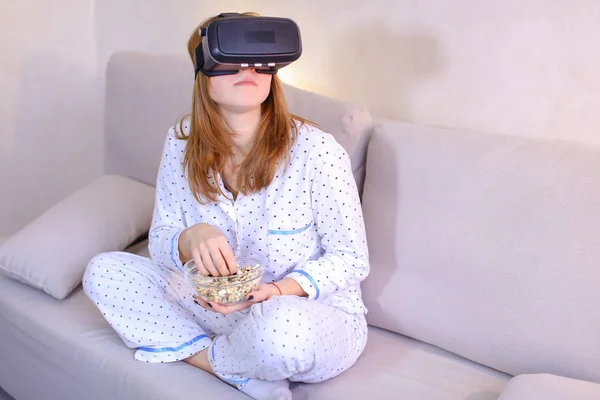 Cool girl watches video in VR glasses, sitting on couch in brigh