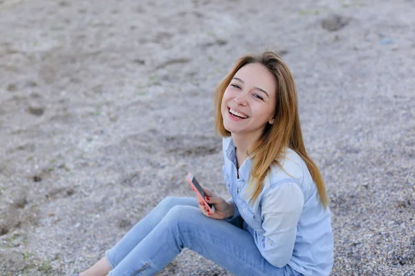 Härlig flicka chattare på telefon med leende och sitter på stranden n — Stockfoto