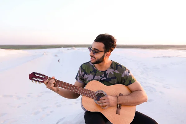 Cute young male Arab enjoys sound of guitar, sitting on hill in