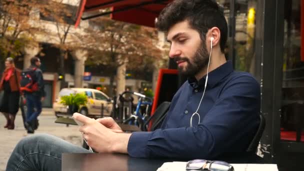 Chico hablando con su novia por teléfono inteligente videollamada y auriculares en la cafetería . — Vídeo de stock