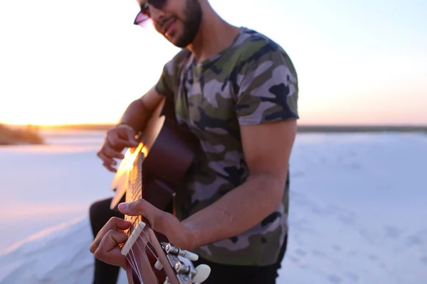 Handsome Arabian guy playing guitar, standing on hill among sand