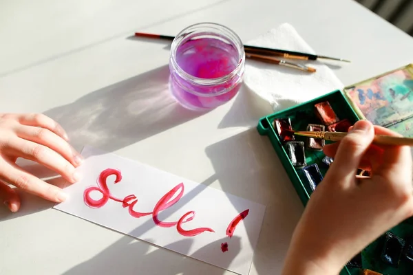Chica pintor escribe con pincel y pinturas inscripción en hoja , — Foto de Stock