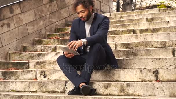 Handsome half Nigerian guy chatting with girl by tablet on steps. — Stock Video